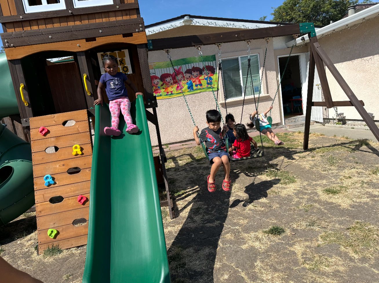 A girl at the top of a green slide while other kids swing